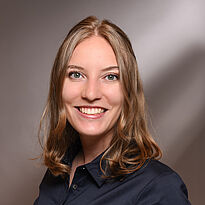 Ein Portrait einer lächelnden Frau mit dunkelblonden, gewellten Haaren, die ein dunkelblaues Hemd trägt. Der Hintergrund ist hell und verschwommen, was die Frau in den Vordergrund hebt.