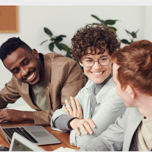Drei Personen sitzen an einem Tisch und lächeln sich an, während sie an Laptops arbeiten. Die Personen haben unterschiedliche Haarfarben und Stilrichtungen und wirken in einem lockeren, kollaborativen Arbeitsumfeld. Pflanzen sind im Hintergrund sichtbar.
