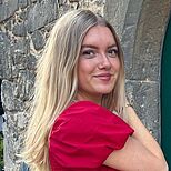 Eine Frau mit langen, blonden Haaren trägt ein rotes Oberteil. Sie steht mit einem leichten Lächeln und zur Seite gedreht, vor einer Steinmauer. Der Hintergrund ist grünlich und unscharf.