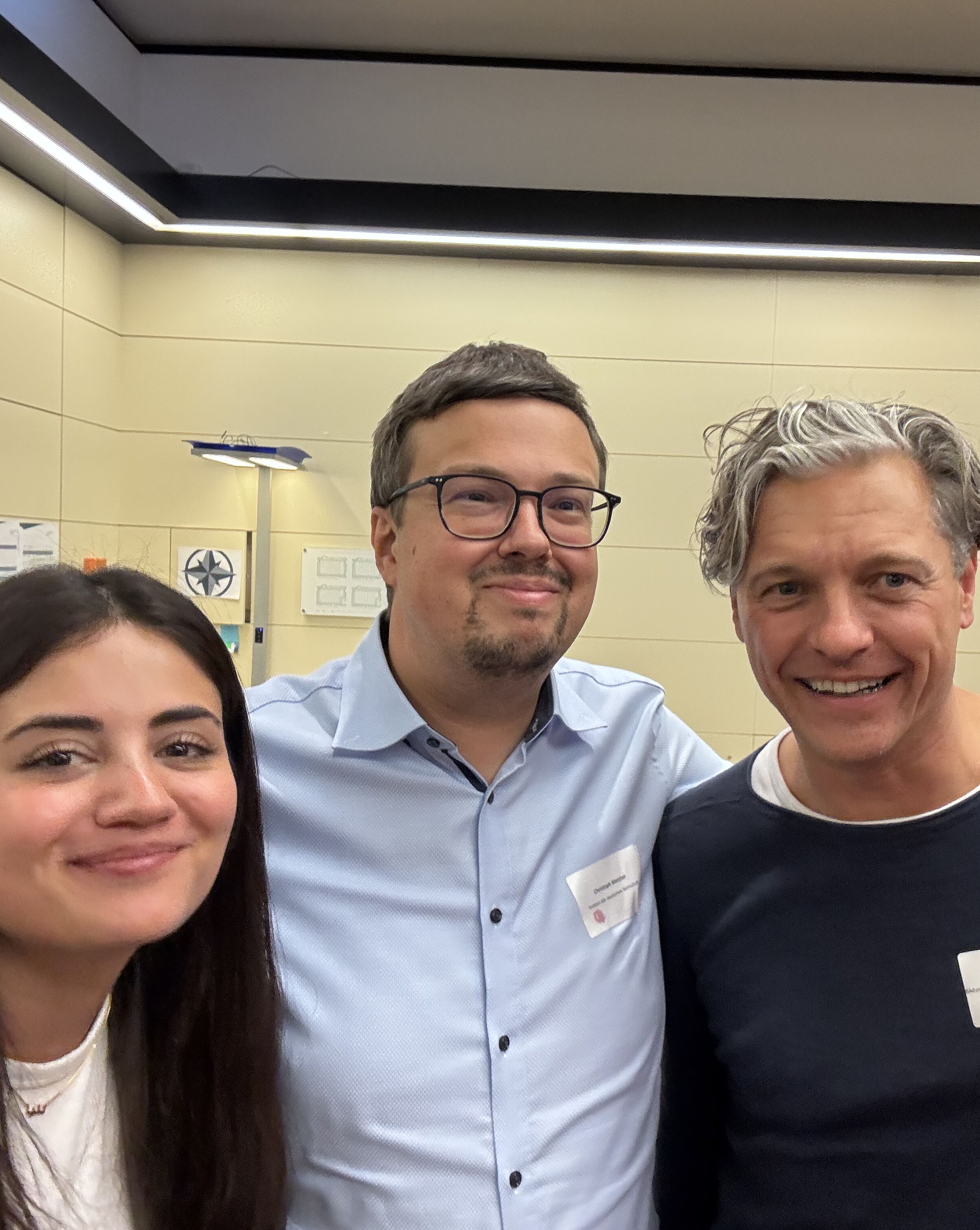 Vier Personen lächeln in die Kamera, während sie sich in einem hellen Raum versammeln. Alle tragen Namensschilder. Die Personen stehen nah beieinander und scheinen ein freundliches Gespräch zu führen.