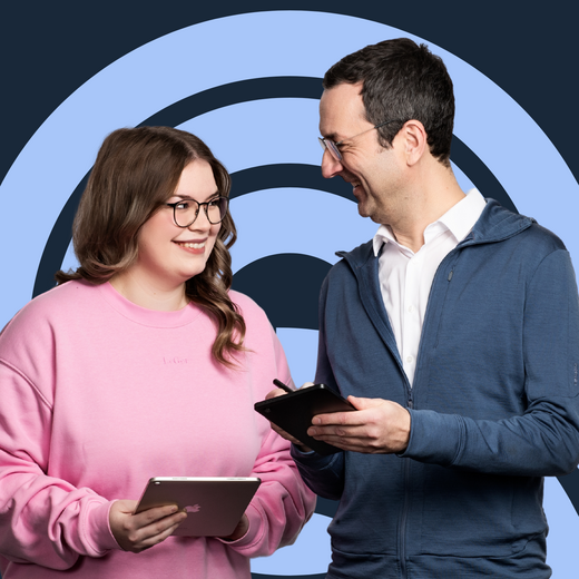 Zwei Personen stehen sich gegenüber und lächeln. Die Frau trägt ein pinkes Sweatshirt und hält ein Tablet, während der Mann in einem blauen Pullover ein Notizbuch in der Hand hat. Im Hintergrund ist ein blauer, kreisförmiger Hintergrund zu sehen.