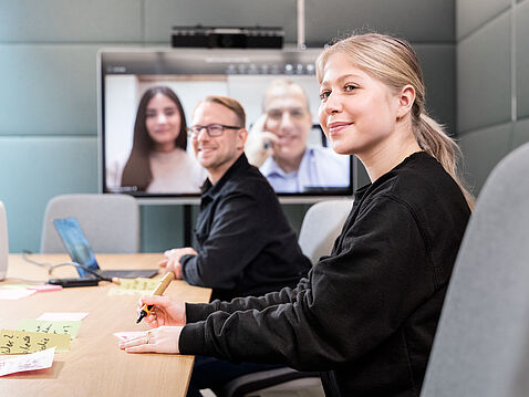 Ein Teammeeting in einem modernen Konferenzraum. Zwei Personen sitzen am Tisch, während sie an einem Videoanruf teilnehmen. Eine Frau schreibt mit einem Stift, ein Mann lächelt. Auf dem Bildschirm sind zwei weitere Personen zu sehen, die an dem Gespräch teilnehmen.