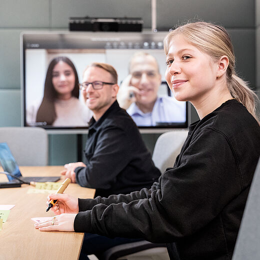 Ein Teammeeting in einem modernen Konferenzraum. Zwei Personen sitzen am Tisch, während sie an einem Videoanruf teilnehmen. Eine Frau schreibt mit einem Stift, ein Mann lächelt. Auf dem Bildschirm sind zwei weitere Personen zu sehen, die an dem Gespräch teilnehmen.