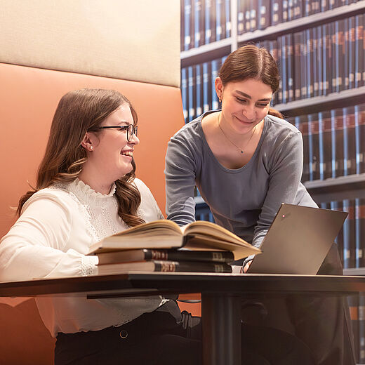 Zwei Frauen sitzen an einem Tisch in einer Bibliothek. Eine Frau lächelt und schaut auf ein geöffnetes Buch, während die andere Frau am Laptop arbeitet und sie anlächelt. Im Hintergrund sind Bücherregale mit vielen Büchern zu sehen.