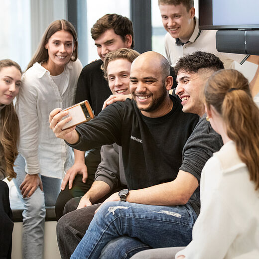 Eine Gruppe von zehn jungen Menschen sitzt zusammen und lächelt. Sie schauen auf ein Smartphone, das eine Person in der Mitte hält. Alle wirken glücklich und vergnügt, während sie diesen gemeinsamen Moment genießen.