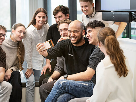Eine Gruppe von zehn jungen Menschen sitzt zusammen und lächelt. Sie schauen auf ein Smartphone, das eine Person in der Mitte hält. Alle wirken glücklich und vergnügt, während sie diesen gemeinsamen Moment genießen.