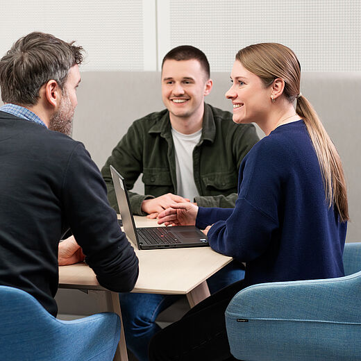 Drei Personen sitzen an einem Tisch in einem modernen Raum, verbunden durch ein Gespräch. Eine Frau zeigt auf den Laptop, während die Männer aufmerksam zuhören und lächeln. Die Atmosphäre wirkt freundlich und kollaborativ.