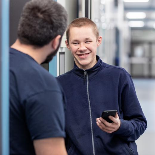 Zwei junge Männer stehen in einem Flur. Der eine, mit einem Smartphone in der Hand, lächelt und schaut den anderen an, der ihm zugewandt ist. Beide tragen Freizeitkleidung und scheinen sich unterhalten zu wollen.