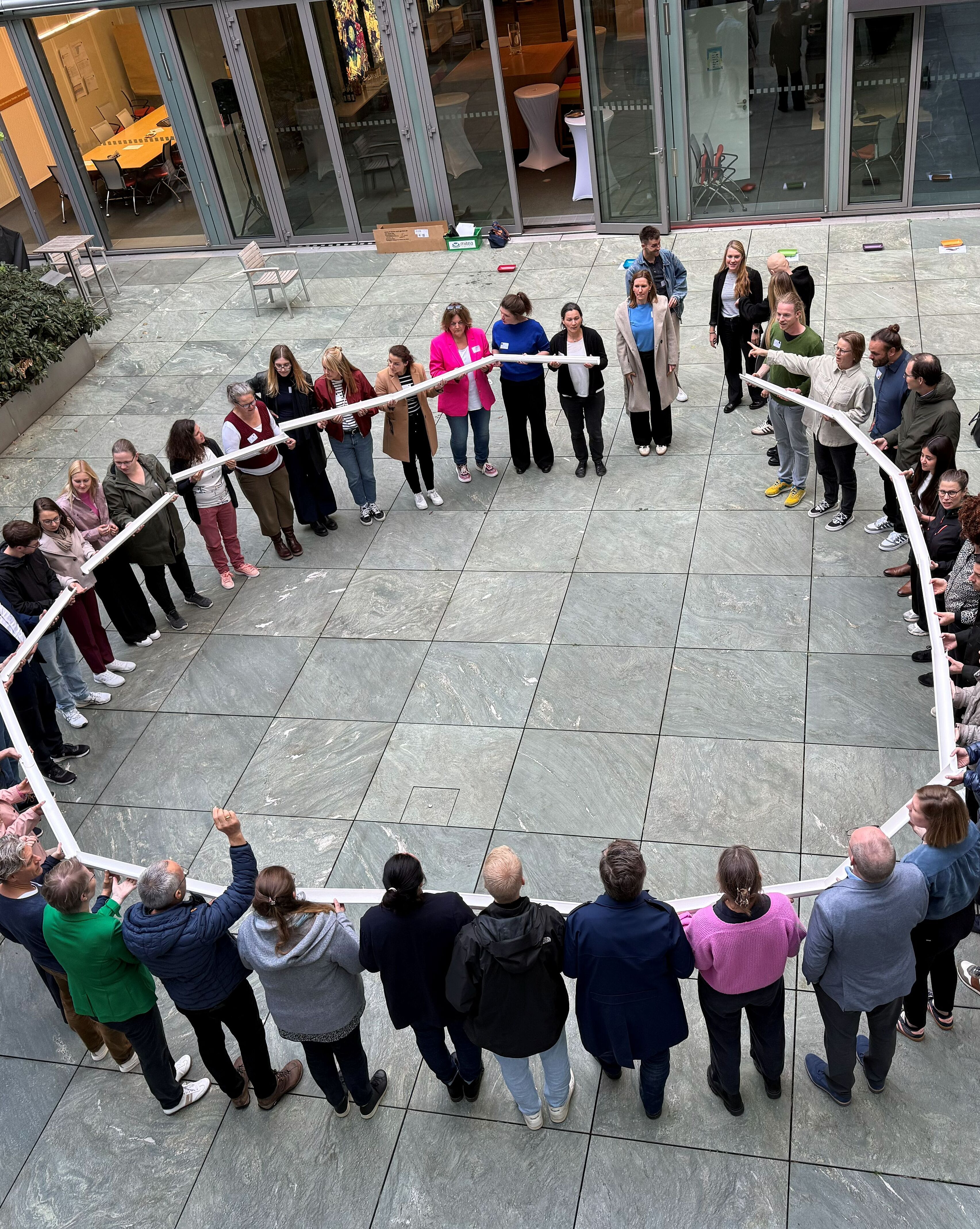 Eine Gruppe von etwa 30 Personen steht in einem Innenhof und bildet einen Kreis. Sie halten ein längliches, weißes Objekt in den Händen. Einige Teilnehmer rufen oder sprechen, während andere aufmerksam zuhören. Der Hintergrund zeigt Pflanzen und moderne Architektur.