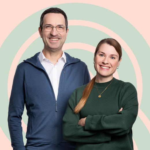 Zwei Personen stehen nebeneinander und lächeln. Der Mann trägt ein blaues Oberteil und eine Brille, die Frau hat einen grünen Pullover und lange Haare. Im Hintergrund sind sanfte, geschwungene Muster in Pastellfarben zu sehen.