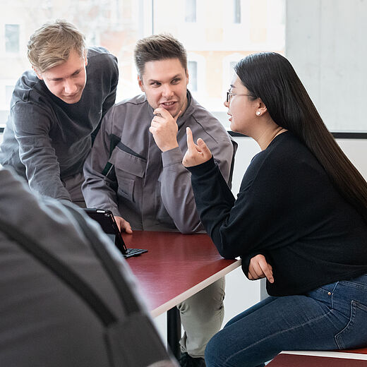 Drei junge Menschen sitzen an einem Tisch und unterhalten sich lebhaft. Zwei Männer schauen auf ein Gerät, während eine Frau gestikuliert und spricht. Der Hintergrund zeigt große Fenster und eine moderne Innenarchitektur.