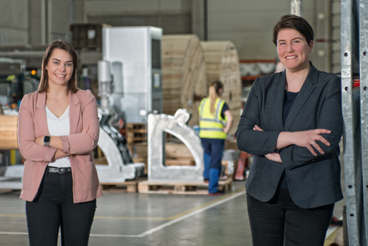 Zwei weibliche Fachkräfte stehen lächelnd in einer Werkshalle. Die eine trägt ein helles Blazer, die andere einen dunklen Anzug. Im Hintergrund sind Maschinen und Mitarbeiterinnen in Arbeitskleidung zu sehen. Die Szene vermittelt eine positive Arbeitsatmosphäre.