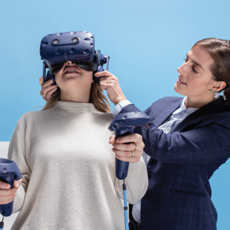Eine Frau trägt ein Virtual-Reality-Headset und hält zwei Steuerungen in den Händen. Eine andere Frau hilft ihr, das Headset korrekt anzupassen. Der Hintergrund ist blau und die Szene vermittelt den Eindruck von interaktiver Technologie.