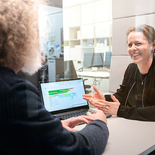 Zwei Personen sitzen an einem Tisch in einem hellen Raum. Eine Frau mit kurzen, blondierten Haaren lächelt und spricht gestikulierend, während ein Mann mit lockigem Haar aufmerksam zuhört. Auf dem Tisch steht ein Laptop, auf dessen Bildschirm eine Tabelle sichtbar ist.