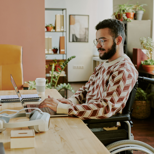 Ein junger Mann im Rollstuhl sitzt an einem Schreibtisch und arbeitet an einem Laptop. Er trägt einen gemusterten Pullover und schaut konzentriert auf den Bildschirm. Im Hintergrund sind Pflanzen und Büromöbel zu sehen.