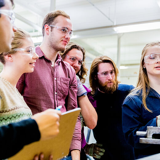Sechs Personen stehen um eine Maschine in einem Werkstattumfeld. Alle tragen Schutzbrillen und sind in einem aufmerksam diskutierenden Austausch. Eine Person erklärt etwas, während die anderen konzentriert zuhören und Notizen machen. Es scheint sich um eine praktische Lern- oder Ausbildungssituation zu handeln.