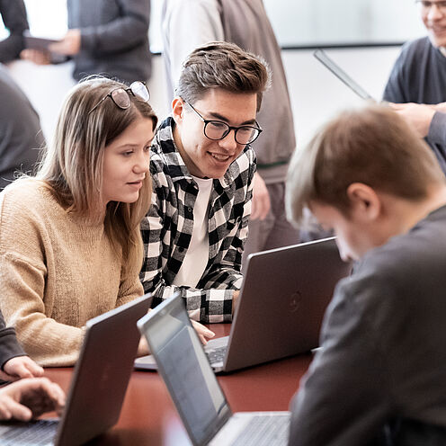 Eine Gruppe von jungen Menschen arbeitet gemeinsam an Laptops. Zwei Personen, ein Mann und eine Frau, schauen interessiert auf den Bildschirm eines Computers. Hinter ihnen sind weitere Personen zu sehen, die an ihren eigenen Laptops arbeiten.