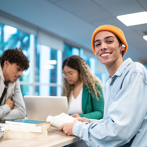 Drei junge Menschen sitzen an einem Tisch. Ein Junge mit orangefarbener Mütze lächelt in die Kamera und hält ein Buch. Im Hintergrund arbeitet ein Mädchen am Laptop, während ein weiterer Junge nachdenklich am Tisch sitzt.