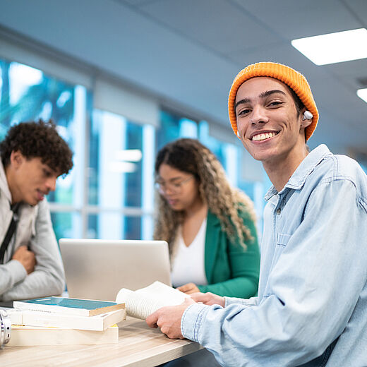 Drei junge Menschen sitzen an einem Tisch. Ein Junge mit orangefarbener Mütze lächelt in die Kamera und hält ein Buch. Im Hintergrund arbeitet ein Mädchen am Laptop, während ein weiterer Junge nachdenklich am Tisch sitzt.