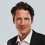 Ein Mann mit lockigem, dunkelblondem Haar lächelt freundlich in die Kamera. Er trägt ein schwarzes Sakko und ein weißes Hemd. Der Hintergrund ist einfarbig hellgrau, wodurch sein Gesicht klar hervorsticht.
