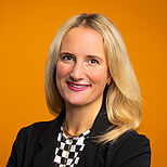 Ein Porträt einer lächelnden Frau mit langen, blonden Haaren. Sie trägt einen schwarzen Blazer und ein gemustertes Oberteil. Der Hintergrund ist orange, was einen freundlichen und einladenden Eindruck vermittelt.