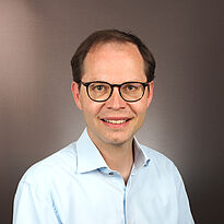 Ein lächelnder Mann mit Brille und kurzen Haaren trägt ein hellblaues Hemd. Der Hintergrund ist einfarbig und neutral.