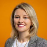 Ein lächelnde Frau mit blonden Haaren trägt ein kariertes Sakko und ein weißes T-Shirt. Der Hintergrund ist orangefarbig, was ein freundliches und einladendes Ambiente schafft.