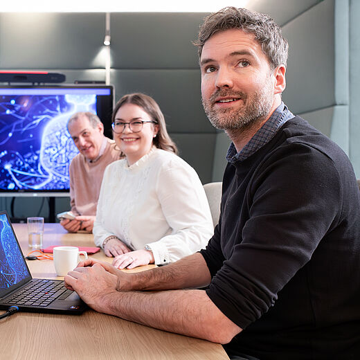 Eine Gruppe von vier Personen sitzt an einem Besprechungstisch. Ein Mann mit Bart schaut zur Kamera. Neben ihm sitzt eine Frau mit Brille. Im Hintergrund sind ein Laptop und ein Bildschirm mit grafischen Inhalten sichtbar. Alle wirken konzentriert und in ein Gespräch vertieft.