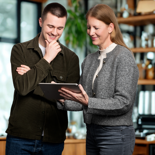 Zwei Personen stehen in einem modernen, holzverzierten Raum und betrachten gemeinsam ein Tablet. Der Mann lächelt und scheint interessiert, während die Frau ihm etwas erklärt. Beide zeigen eine positive, engagierte Interaktion.