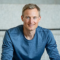 Ein lächelnder Mann mit kurzem, blondem Haar trägt ein blaues Oberteil. Er sitzt vor einer grauen, strukturierten Wand. Die Bildkomposition strahlt Freundlichkeit und Selbstbewusstsein aus.