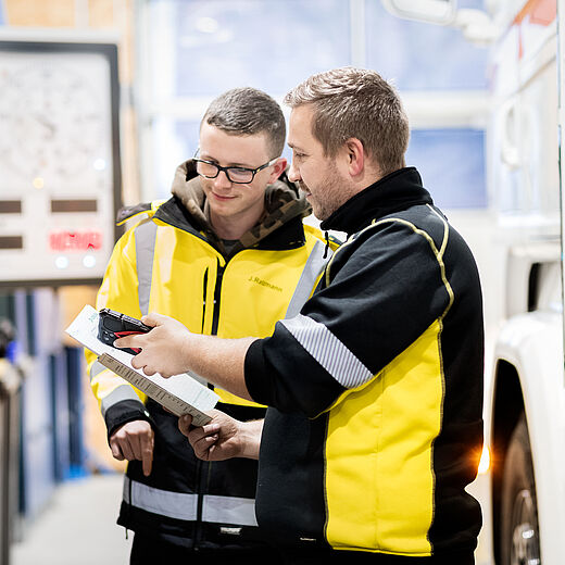 Zwei Männer in gelben Arbeitsjacken stehen in einer Werkstatt. Sie schauen auf ein Dokument, während einer von ihnen etwas erklärt. Der Hintergrund zeigt technische Geräte und einen Lkw, was auf eine technische oder handwerkliche Umgebung hinweist.