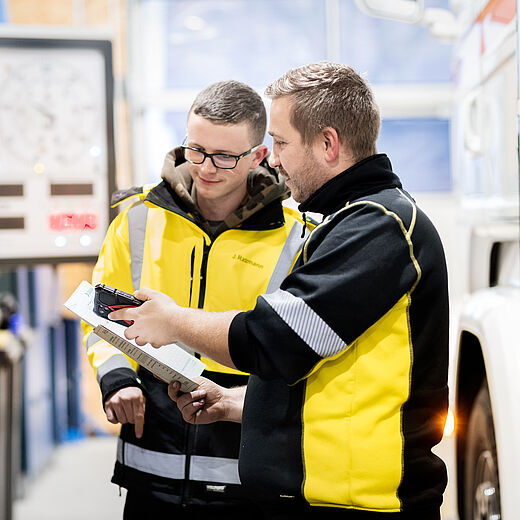 Zwei Männer in gelben Arbeitsjacken stehen in einer Werkstatt. Sie schauen auf ein Dokument, während einer von ihnen etwas erklärt. Der Hintergrund zeigt technische Geräte und einen Lkw, was auf eine technische oder handwerkliche Umgebung hinweist.