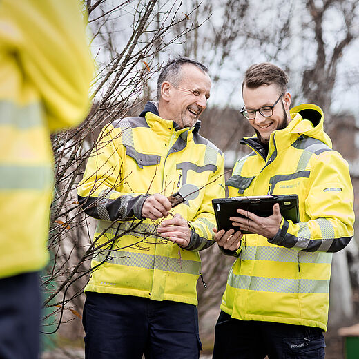 Zwei Männer in gelben Sicherheitsjacken stehen draußen. Einer hält eine Zange und lächelt, während der andere ein Tablet in der Hand hält und auf den Bildschirm schaut. Im Hintergrund sind kahle Bäume zu sehen.