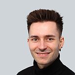 Ein junger Mann mit kurzen, gestuften Haaren lächelt freundlich in die Kamera. Er trägt einen schwarzen Rollkragenpullover. Der Hintergrund ist ein einfarbiger grauer Farbton.