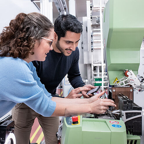 Zwei Personen, eine Frau mit Locken und Brille sowie ein Mann, stehen an einer Maschine. Sie arbeiten zusammen, während sie Teile der Maschine ansehen und bedienen. Der Fokus liegt auf ihrer gemeinsamen Aktivität und dem technischen Gerät.