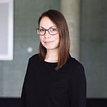 Eine lächelnde Frau mit langen, braunen Haaren und einer schwarzen Bluse steht vor einem modernen Hintergrund. Sie trägt eine modische Brille und hat einen freundlichen Ausdruck.