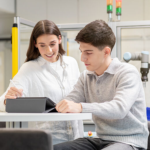 Zwei junge Menschen arbeiten zusammen an einem Tisch. Eine Frau erklärt etwas und zeigt auf ein Gerät, während ein Mann aufmerksam zuhört und Notizen macht. Im Hintergrund sind Technik und Arbeitsmaterialien sichtbar.