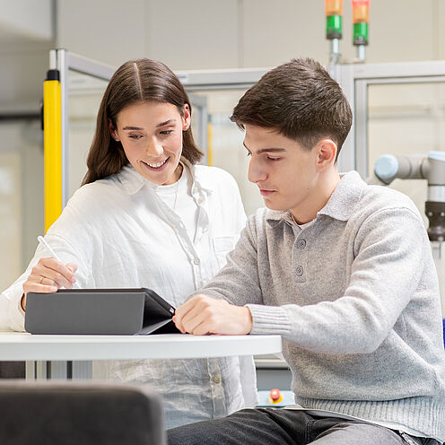Zwei junge Menschen arbeiten zusammen an einem Tisch. Eine Frau erklärt etwas und zeigt auf ein Gerät, während ein Mann aufmerksam zuhört und Notizen macht. Im Hintergrund sind Technik und Arbeitsmaterialien sichtbar.