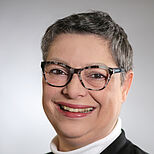 Porträt einer lächelnden Frau mit kurzen grauen Haaren, die eine Brille trägt. Sie hat einen weißen Rollkragenpullover und eine dunkle Jacke an. Der Hintergrund ist hell und unifarben.