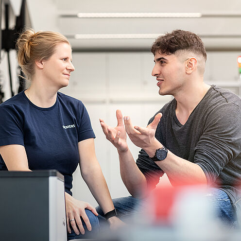 Zwei Personen führen ein Gespräch. Eine Frau mit blonden Haaren trägt ein dunkelblaues Shirt, während ein Mann mit kurzen, braunen Haaren und einem grauen Pullover lebhaft gestikuliert. Beide sitzen in einem modernen, hellen Umfeld.