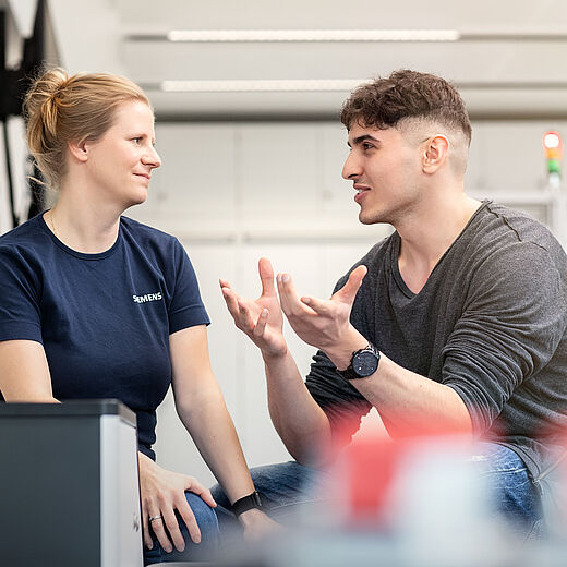Zwei Personen führen ein Gespräch. Eine Frau mit blonden Haaren trägt ein dunkelblaues Shirt, während ein Mann mit kurzen, braunen Haaren und einem grauen Pullover lebhaft gestikuliert. Beide sitzen in einem modernen, hellen Umfeld.