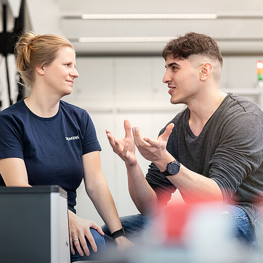 Zwei Personen führen ein Gespräch. Eine Frau mit blonden Haaren trägt ein dunkelblaues Shirt, während ein Mann mit kurzen, braunen Haaren und einem grauen Pullover lebhaft gestikuliert. Beide sitzen in einem modernen, hellen Umfeld.
