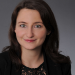 Porträt einer Frau mit braunen Haaren, die in einem schwarzen Blazer gekleidet ist. Sie lächelt freundlich in die Kamera, vor einem grauen Hintergrund. Ihre Augen sind hell und ausdrucksstark.