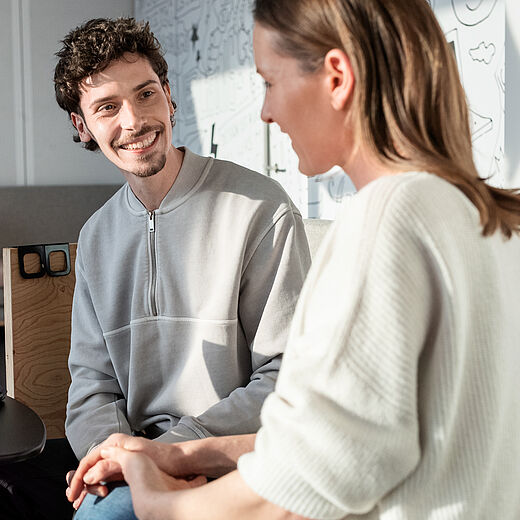 Ein junger Mann und eine junge Frau sitzen in einem modernen Büro-Setup. Der Mann lächelt, während er mit der Frau spricht, die ihm gegenüber sitzt. Ein Laptop steht auf einem Tisch zwischen ihnen, und der Raum ist hell erleuchtet.