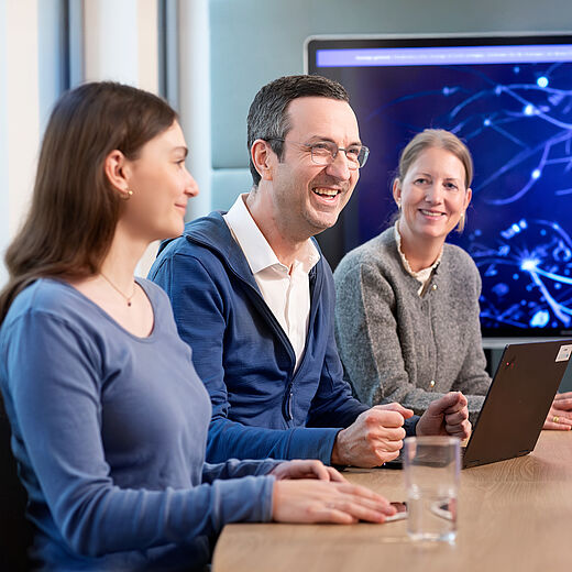 Drei Personen sitzen an einem Konferenztisch und lächeln. Ein Mann in blauer Jacke hält ein Tablet. Die Frauen neben ihm tragen bequeme Kleidung. Im Hintergrund ist ein Bildschirm mit abstrakter Technologie-Darstellung sichtbar.