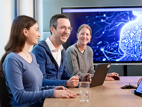 Drei Personen sitzen an einem Konferenztisch und lächeln. Ein Mann in blauer Jacke hält ein Tablet. Die Frauen neben ihm tragen bequeme Kleidung. Im Hintergrund ist ein Bildschirm mit abstrakter Technologie-Darstellung sichtbar.