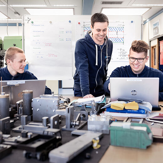 Drei junge Männer in blauen Sweatshirts sitzen an einem Tisch in einem Arbeitsraum. Sie arbeiten gemeinsam am Computer, während technische Bauteile und Werkzeuge vor ihnen ausgebreitet sind. Der Mann in der Mitte lächelt und zeigt auf den Bildschirm.