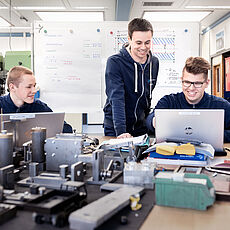Drei junge Männer in blauen Sweatshirts sitzen an einem Tisch in einem Arbeitsraum. Sie arbeiten gemeinsam am Computer, während technische Bauteile und Werkzeuge vor ihnen ausgebreitet sind. Der Mann in der Mitte lächelt und zeigt auf den Bildschirm.