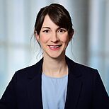 Eine Frau mit dunklem Haar und blauer Bluse trägt einen schwarzen Blazer. Sie lächelt freundlich in die Kamera. Der Hintergrund ist unscharf und hell.