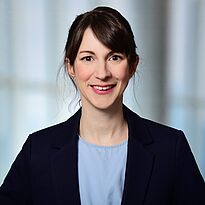 Eine Frau mit dunklem Haar und blauer Bluse trägt einen schwarzen Blazer. Sie lächelt freundlich in die Kamera. Der Hintergrund ist unscharf und hell.